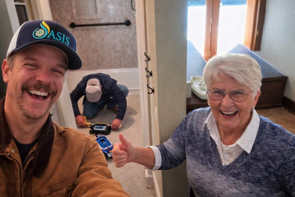 A smiling man in a cap and an older woman give a thumbs-up while a worker installs equipment in a bathroom in the background. Sunlight streams through a window.